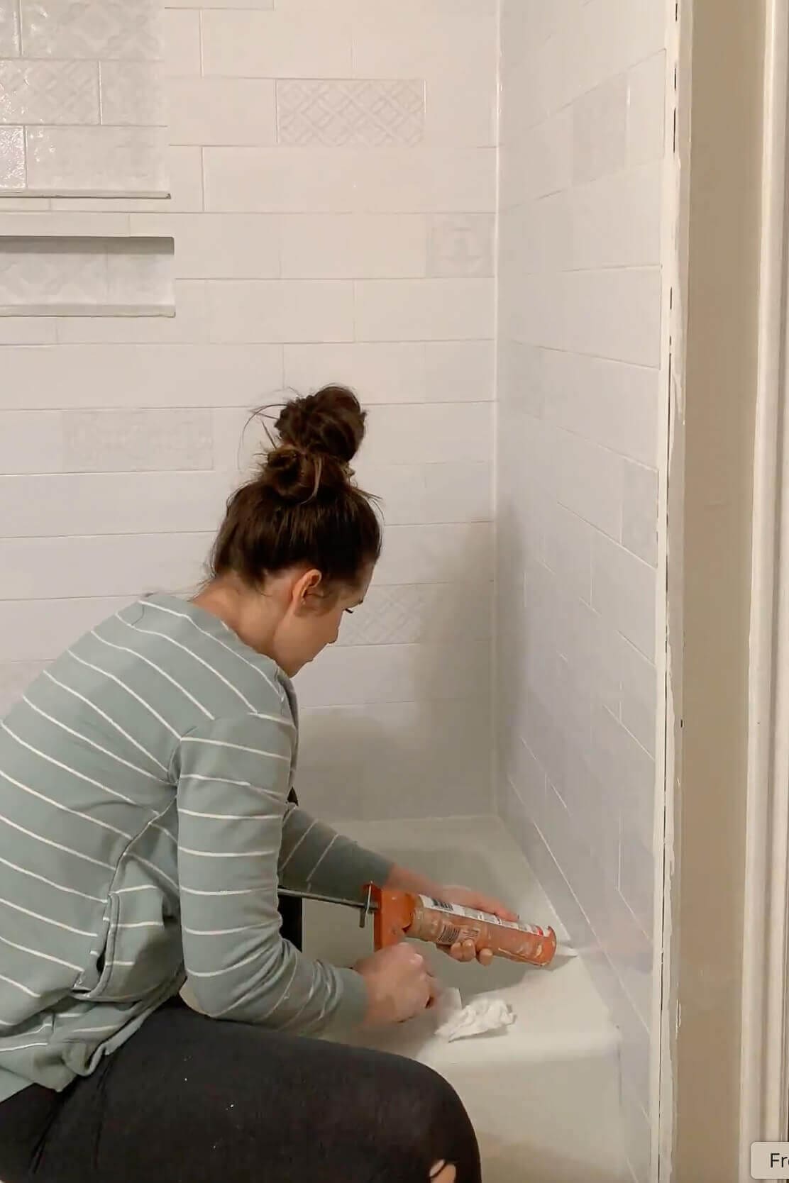 Woman sitting on the tub ledge caulking the seam where the white subway tile shower wall meets the bathtub.