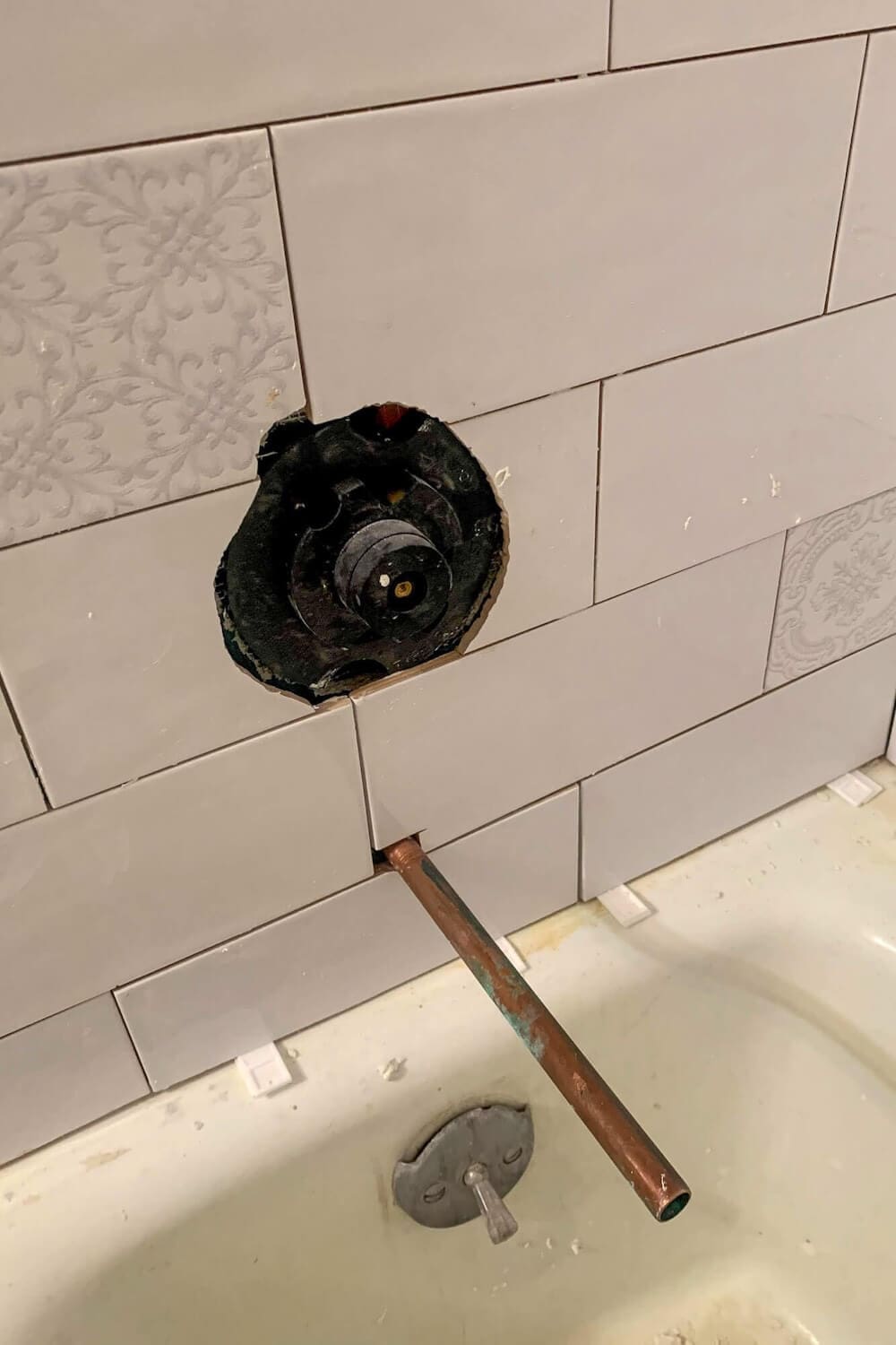 Close-up of tiled shower wall showing exposed black mixing valve and copper tub spout pipe before faucet trim installation.