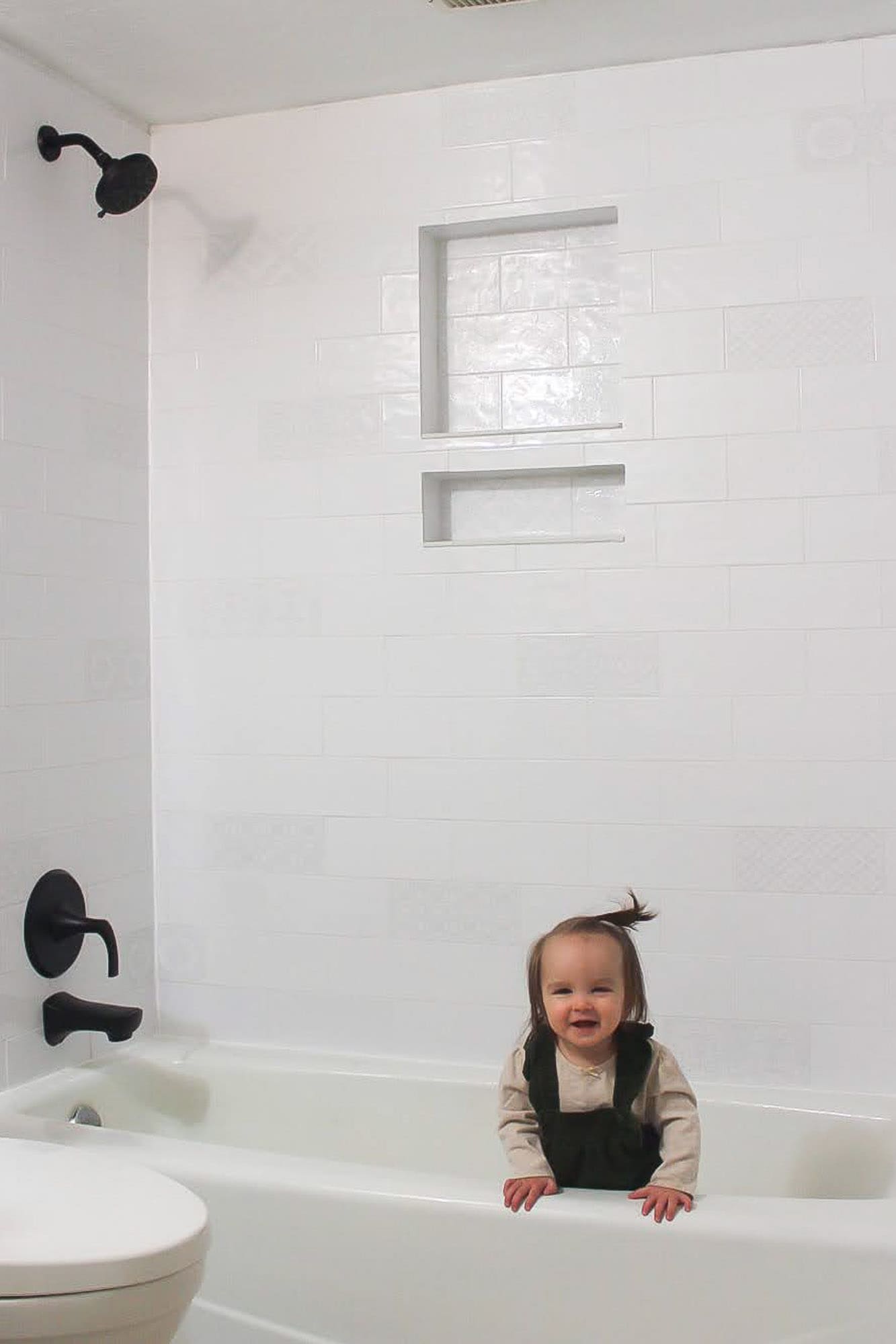 Smiling toddler standing in the bathtub in front of a finished white subway tile shower with double niche and matte black fixtures.