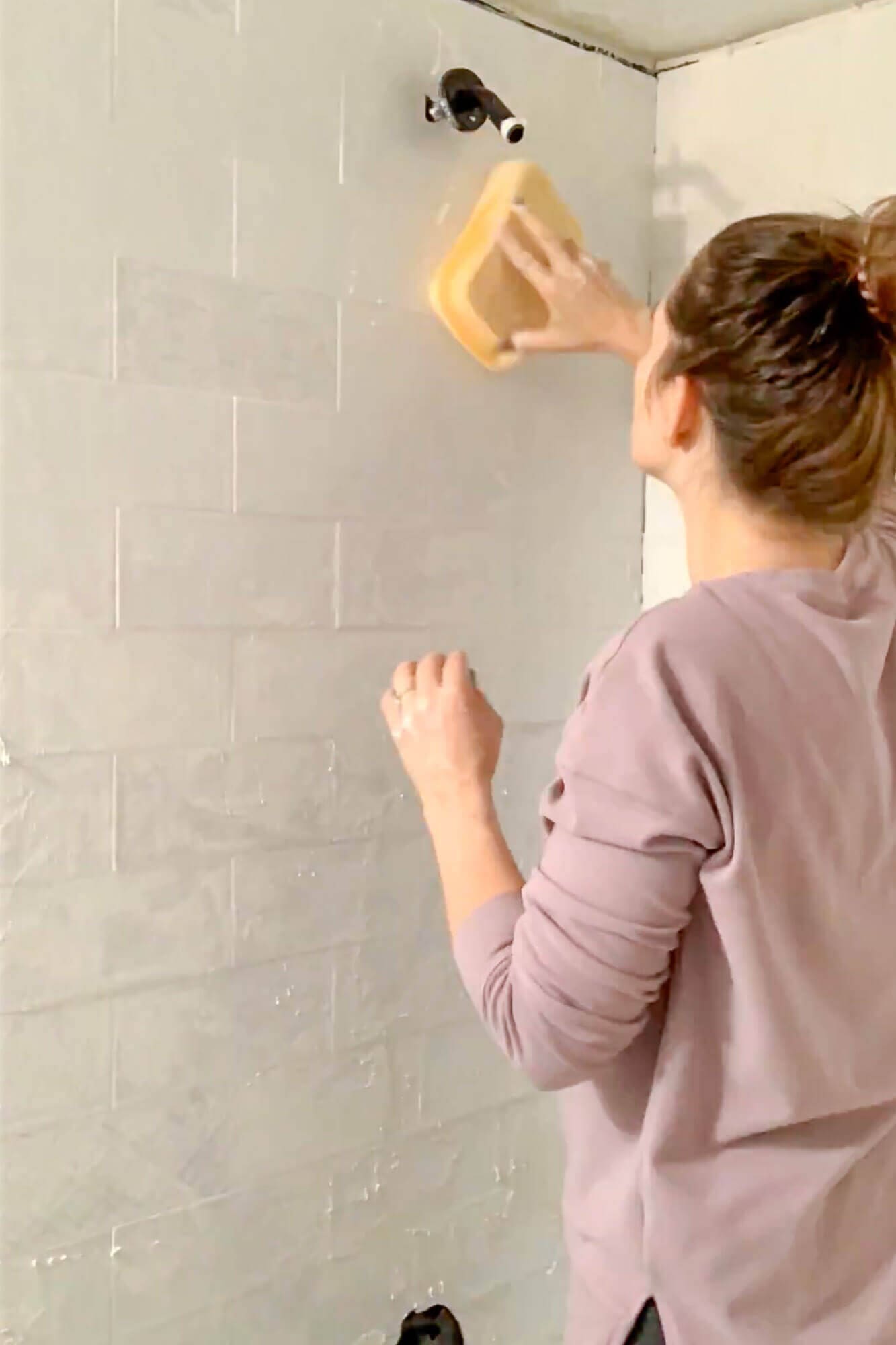Wiping excess grout from shower wall with sponge.