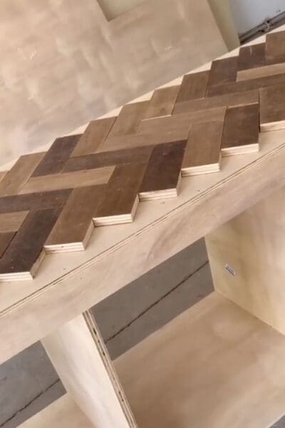 first rows of a wood herringbone pattern laid along the front edge of a plywood cabinet