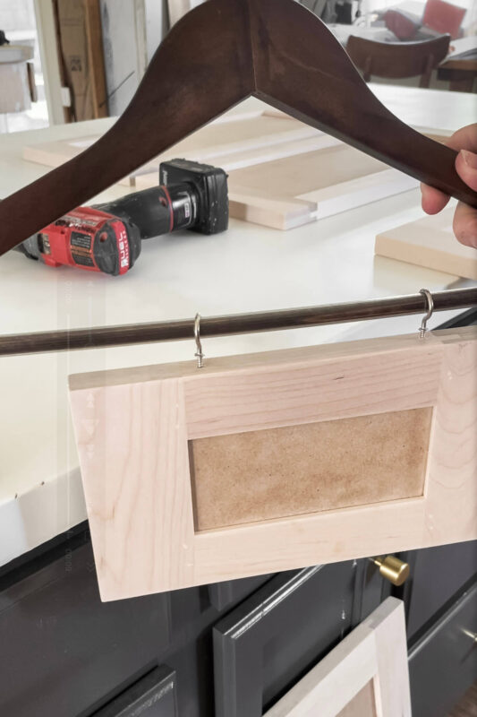 Unfinished cabinet door hanging from a wooden clothes hanger, with a drill and other cabinet parts visible in the background, part of a DIY kitchen remodel.