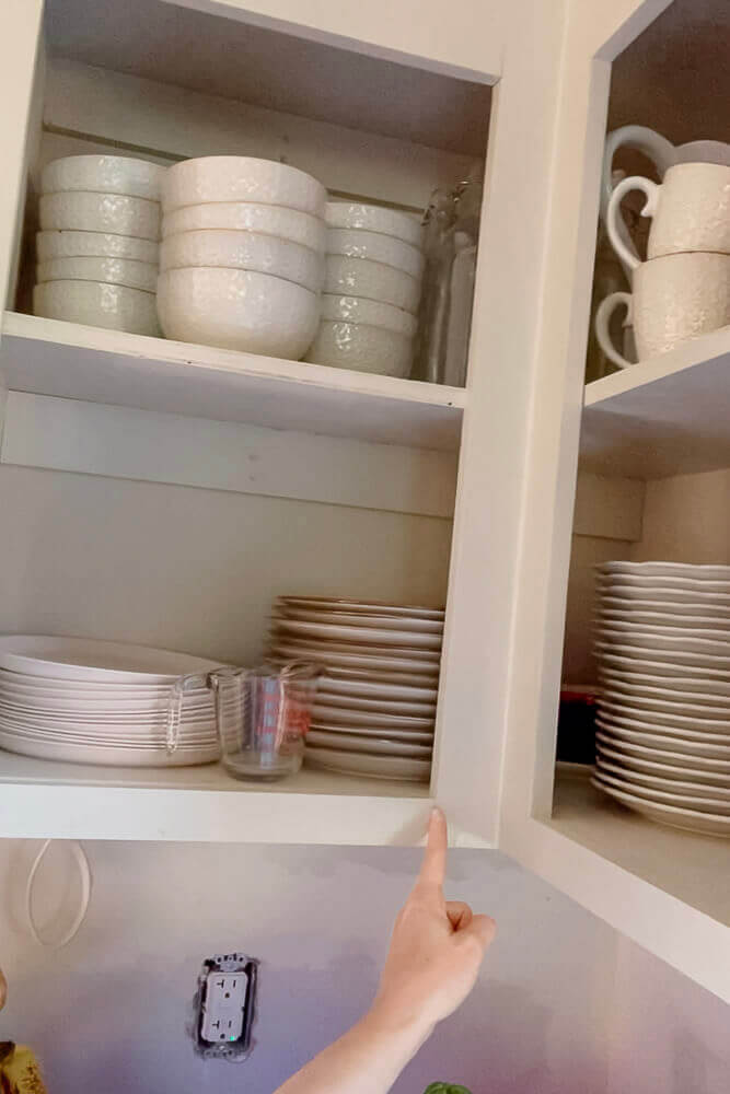 Person pointing to a corner cabinet shelf filled with white dishes, part of an affordable kitchen remodel.