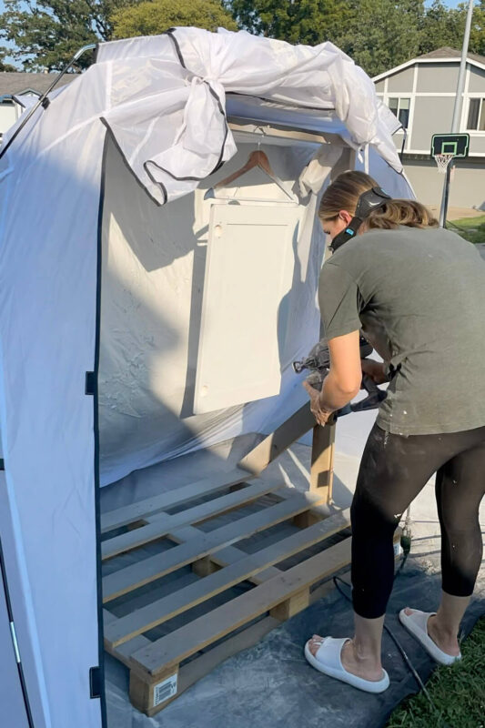 A person wearing a mask sprays white paint onto a cabinet door in an outdoor setup. The door is hanging from a hanger inside a makeshift spray booth made of wood and plastic sheeting.