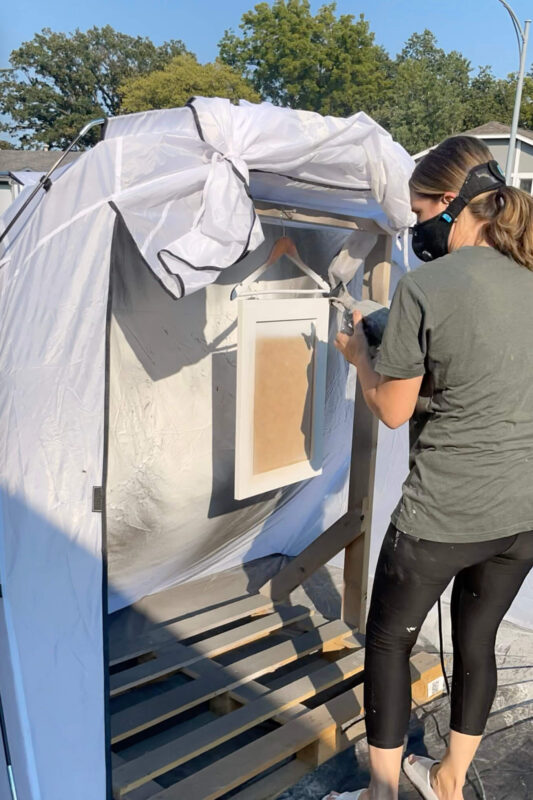 A person wearing a mask sprays white paint onto a cabinet door in an outdoor setup. The door is hanging from a hanger inside a makeshift spray booth made of wood and plastic sheeting.