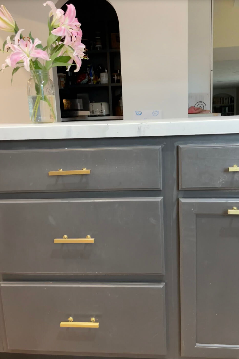 Close-up of dark gray kitchen cabinets with gold handles, topped with a white countertop. A vase of pink lilies sits on the counter, with an arched opening in the background showing a shelf.