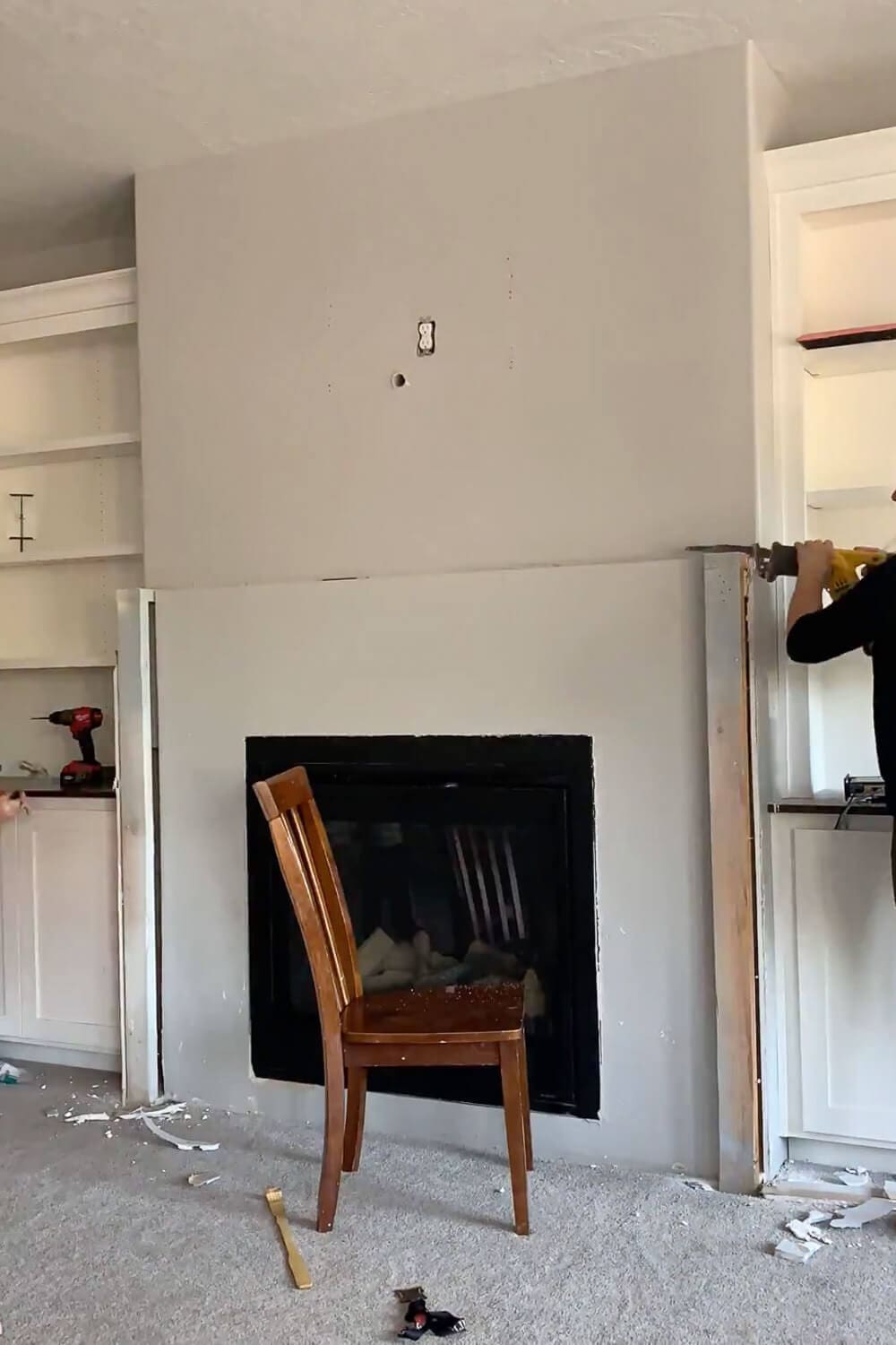 Primed fireplace wall mid-renovation with person using drill on the side, chair in front of fireplace insert, construction debris on floor