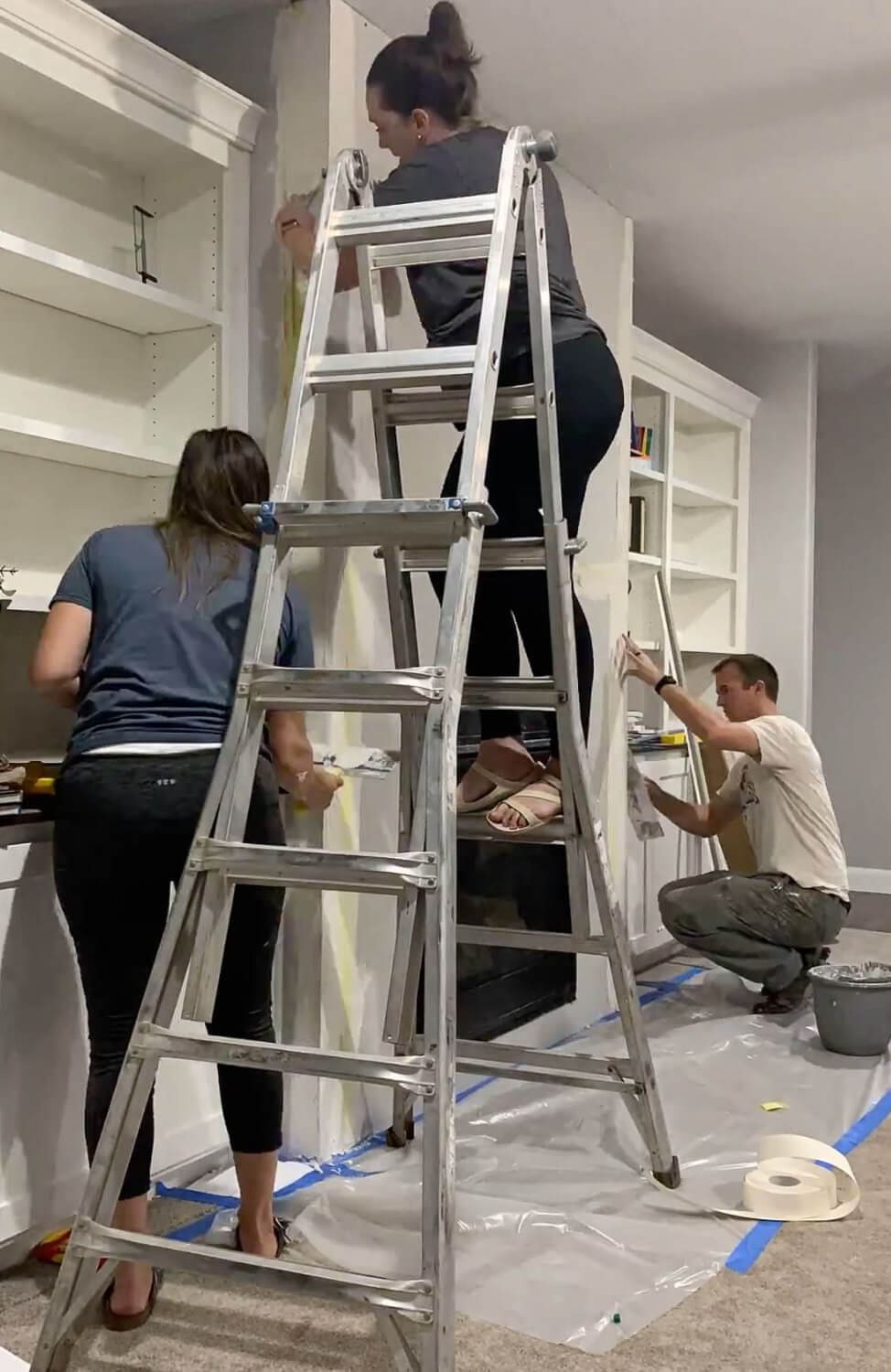 Three people prepping fireplace for renovation - one on ladder, two below taping and positioning, plastic drop cloth on floor