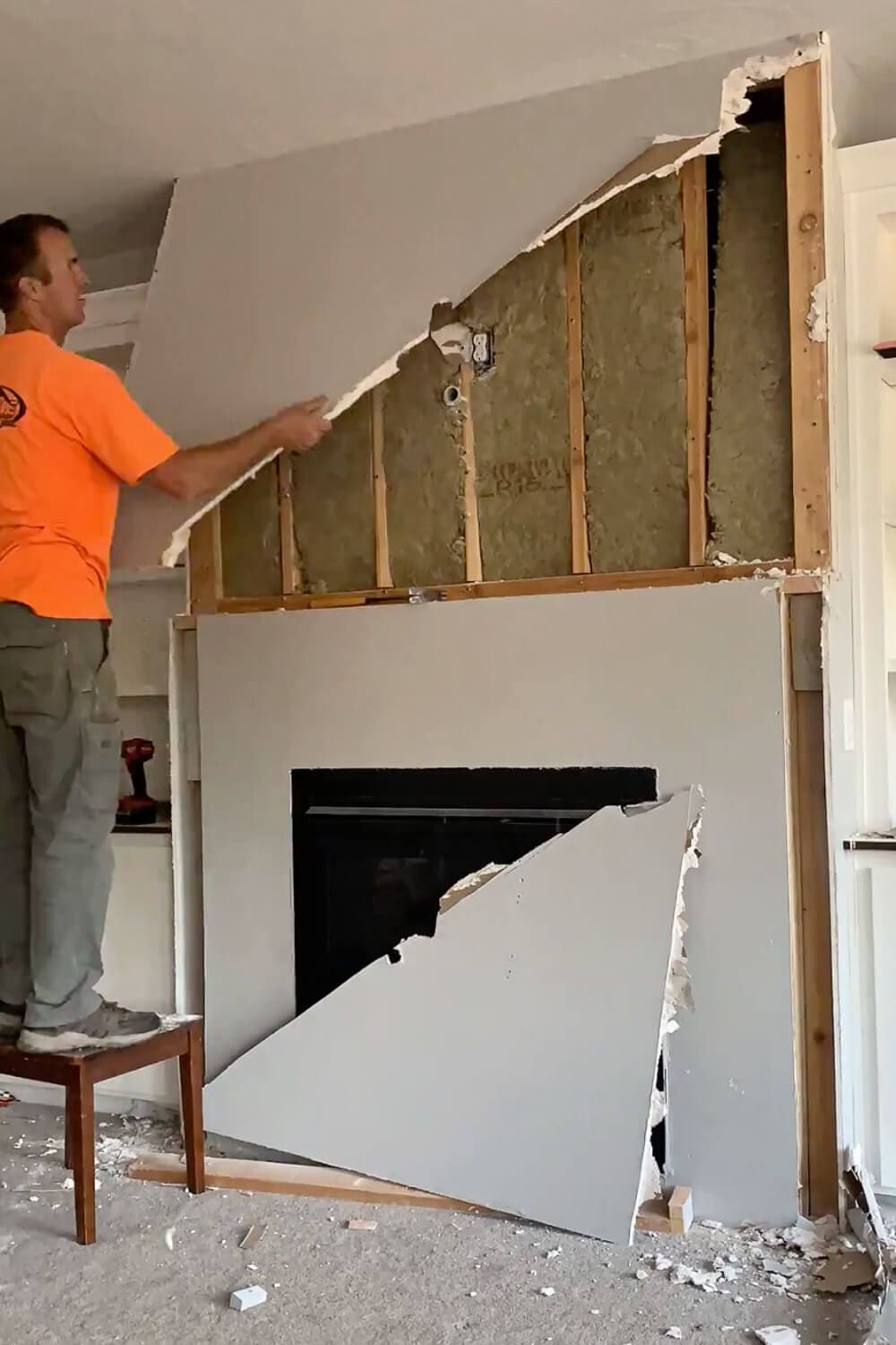 Man in orange shirt pulling down drywall from above fireplace exposing wood studs and insulation, large drywall pieces falling