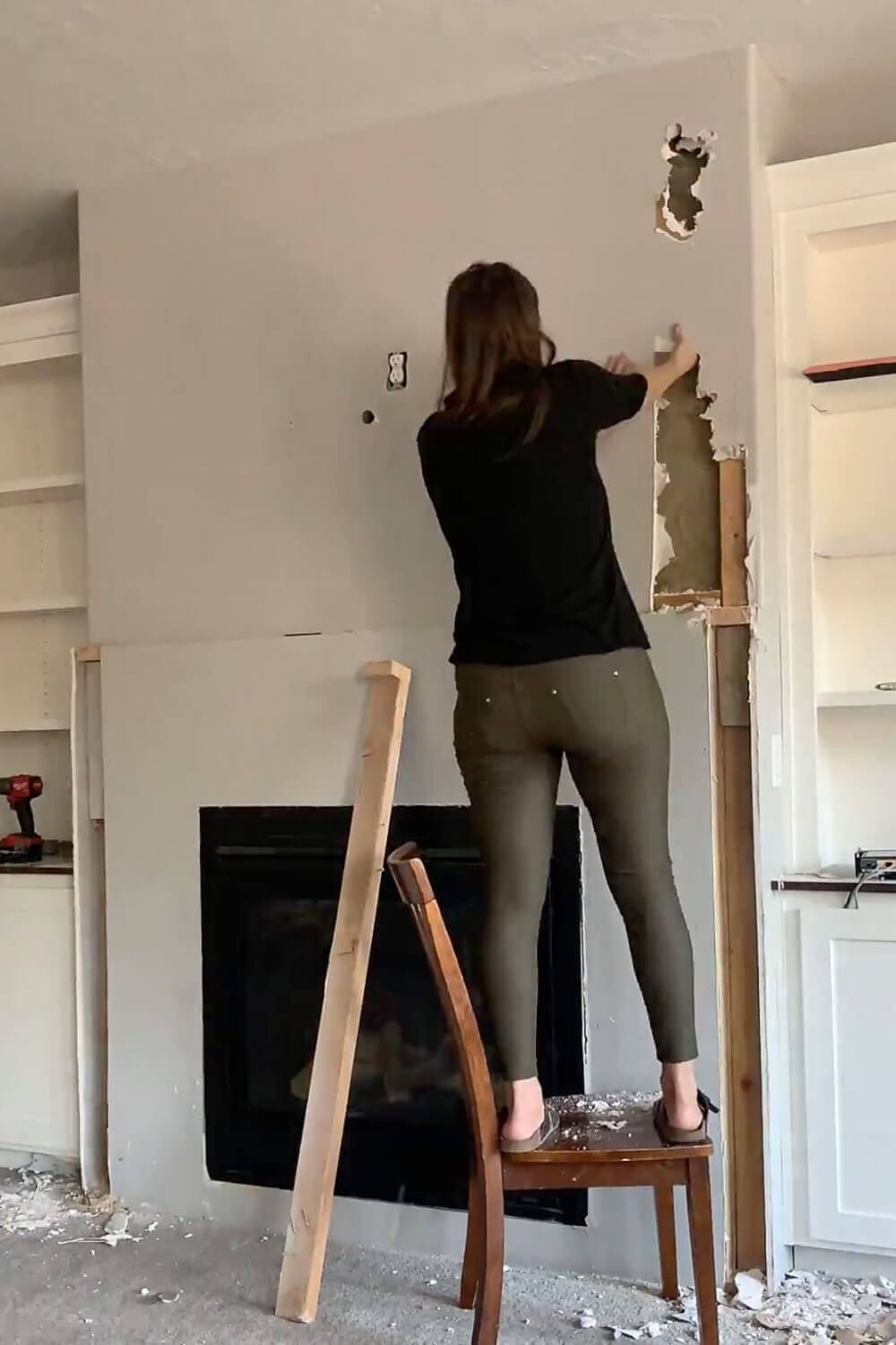 Woman standing on chair tearing drywall from side of fireplace during demolition, exposed wood studs visible, construction debris on floor