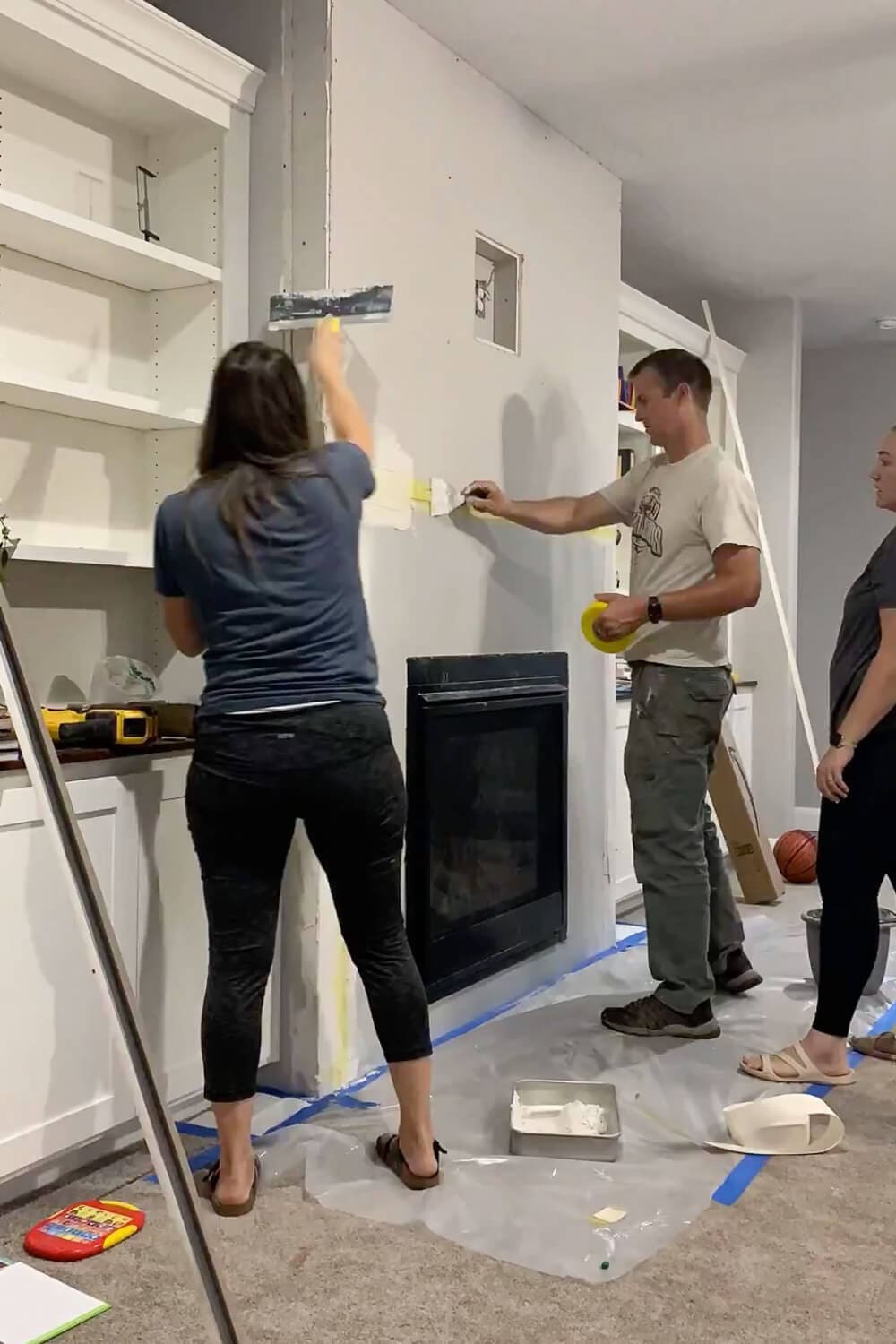 Multiple people applying joint compound to fireplace drywall - woman with taping knife spreading mud while man holds painters tape