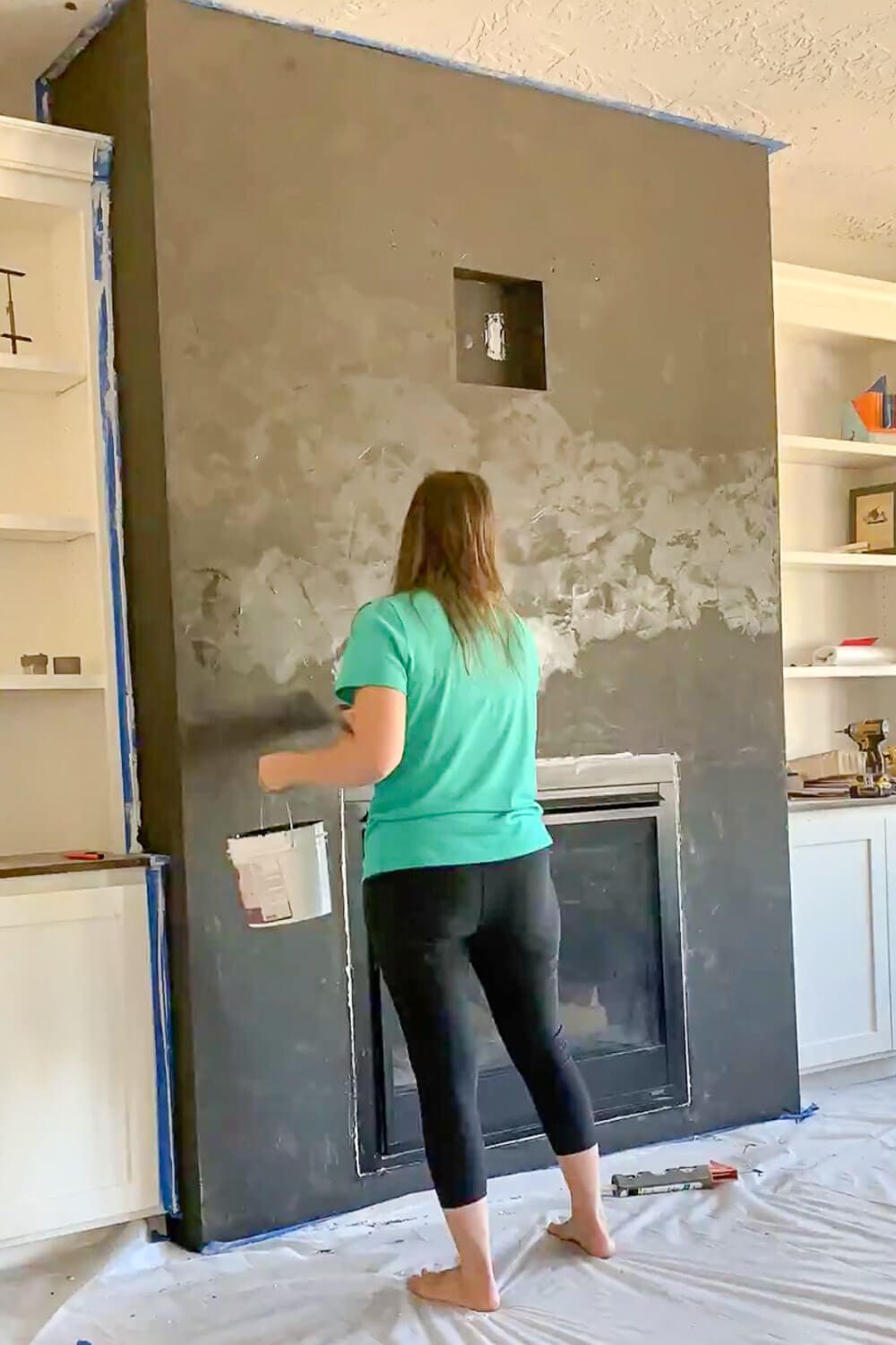 Woman in teal shirt applying second coat of black plaster to fireplace, half the wall covered showing contrast between coats