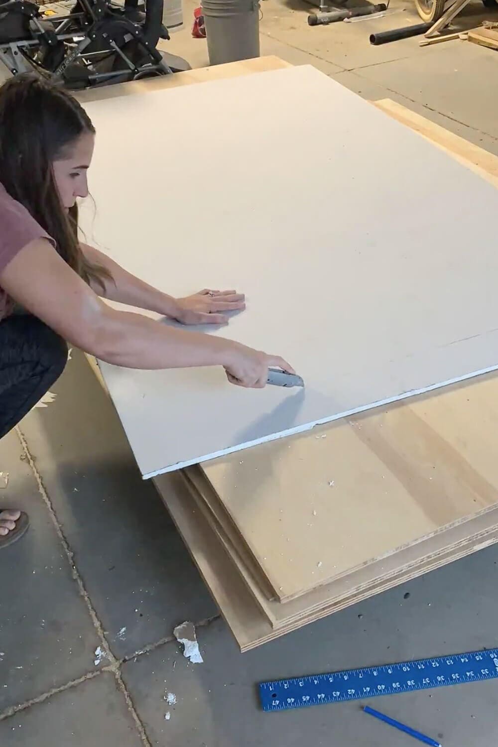 Woman scoring sheetrock with utility knife on plywood in garage, drywall square visible for measuring straight cuts