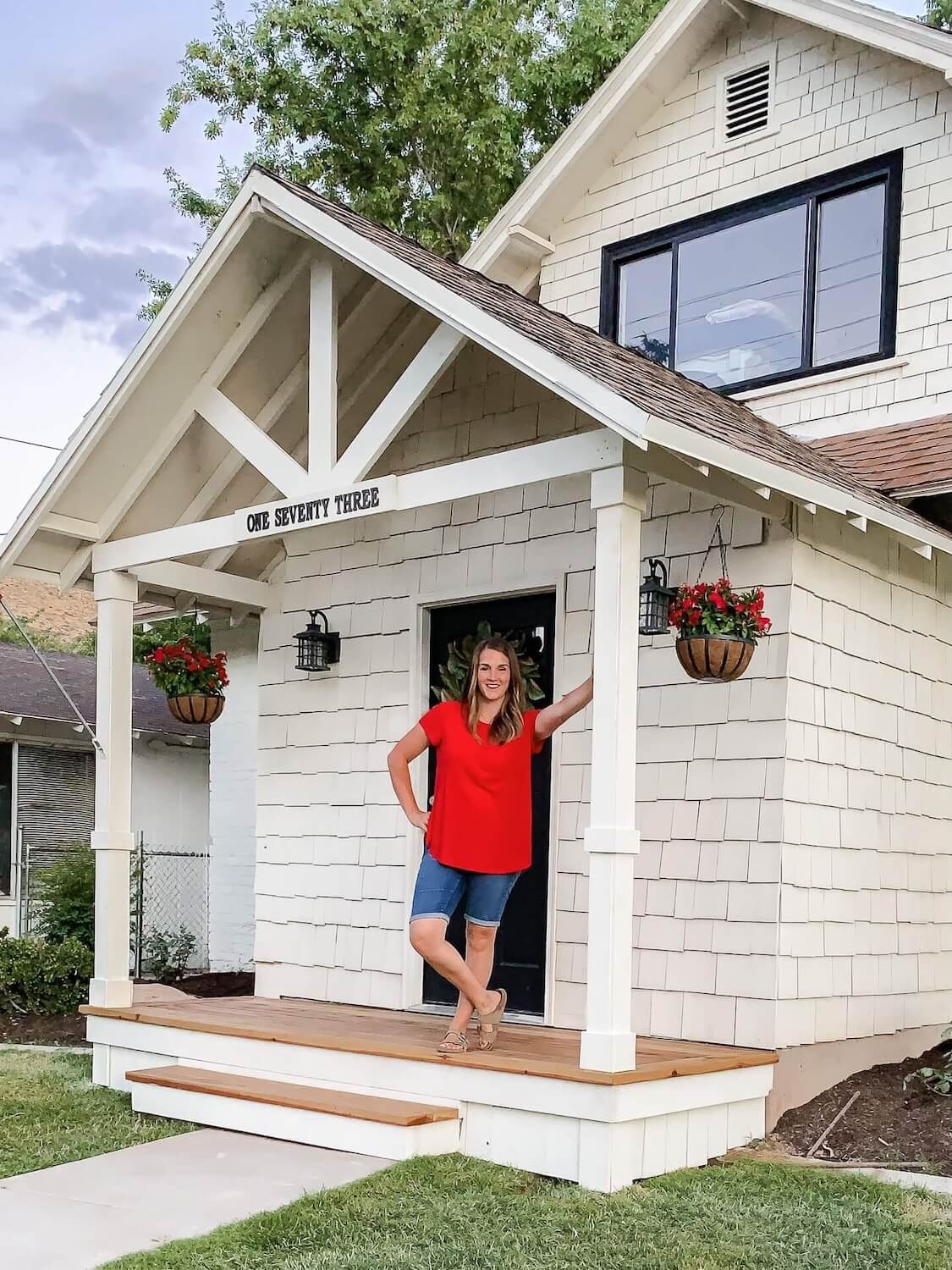Crystel standing on her newly built white front porch, leaning against a column beside the black front door and hanging flower baskets.