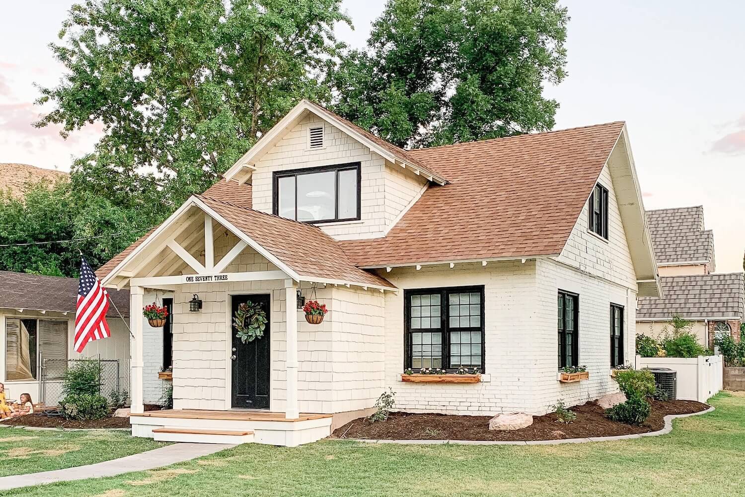 Front view of a craftsman house after adding a front porch to a house, with white columns, an American flag, window flower boxes, and kids playing on the lawn.