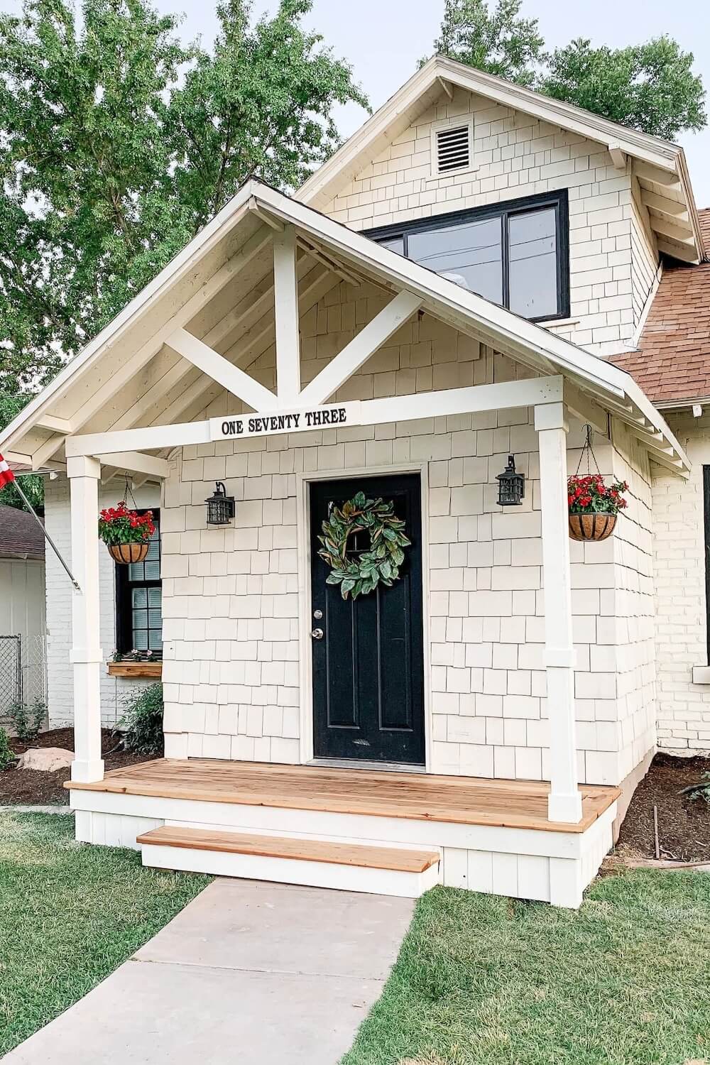 Angled view of a finished white DIY front porch with a black front door, green wreath, hanging baskets of red flowers, and a wood deck.