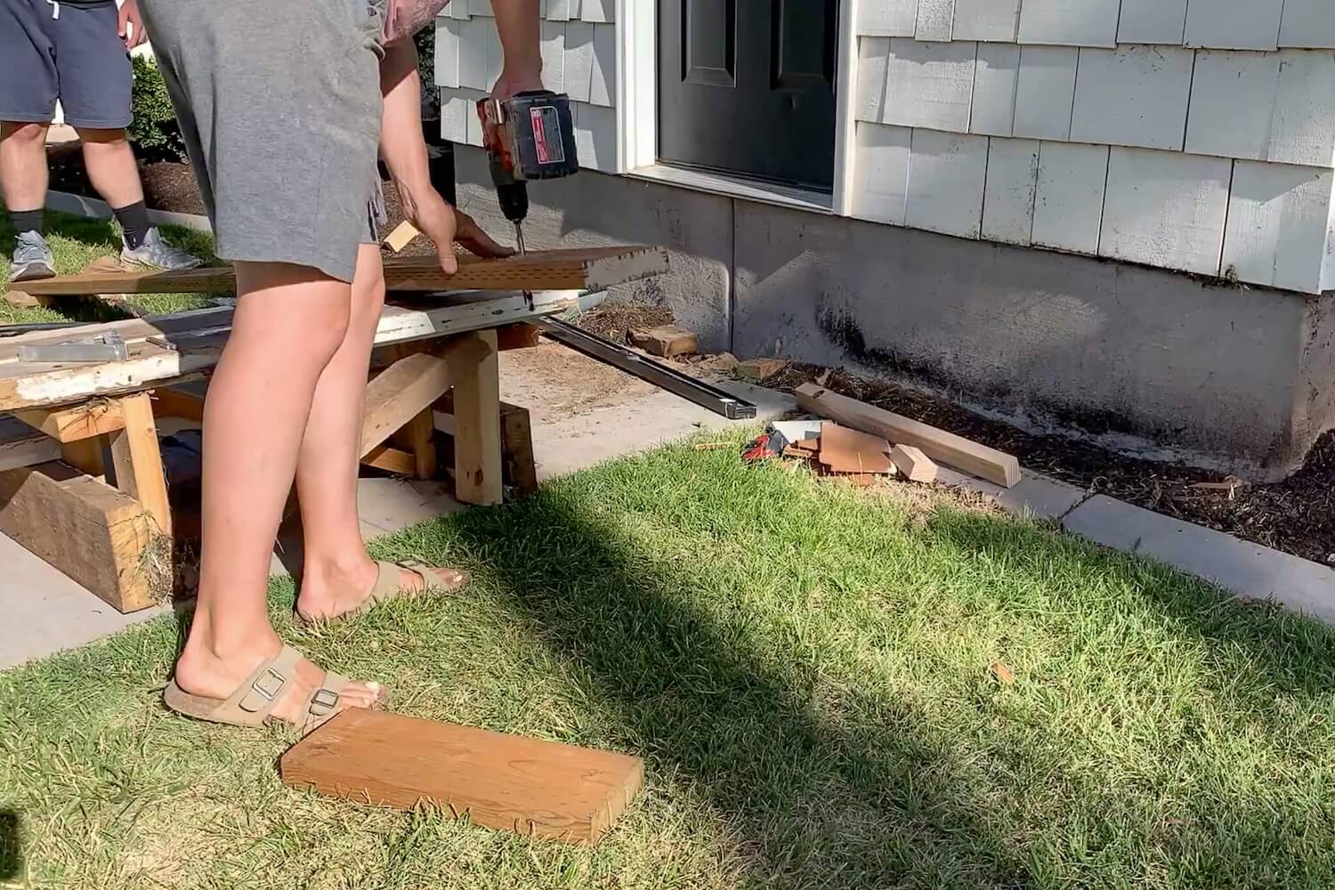 Two people drilling pilot holes into the ledger board against the house siding using a hammer drill.