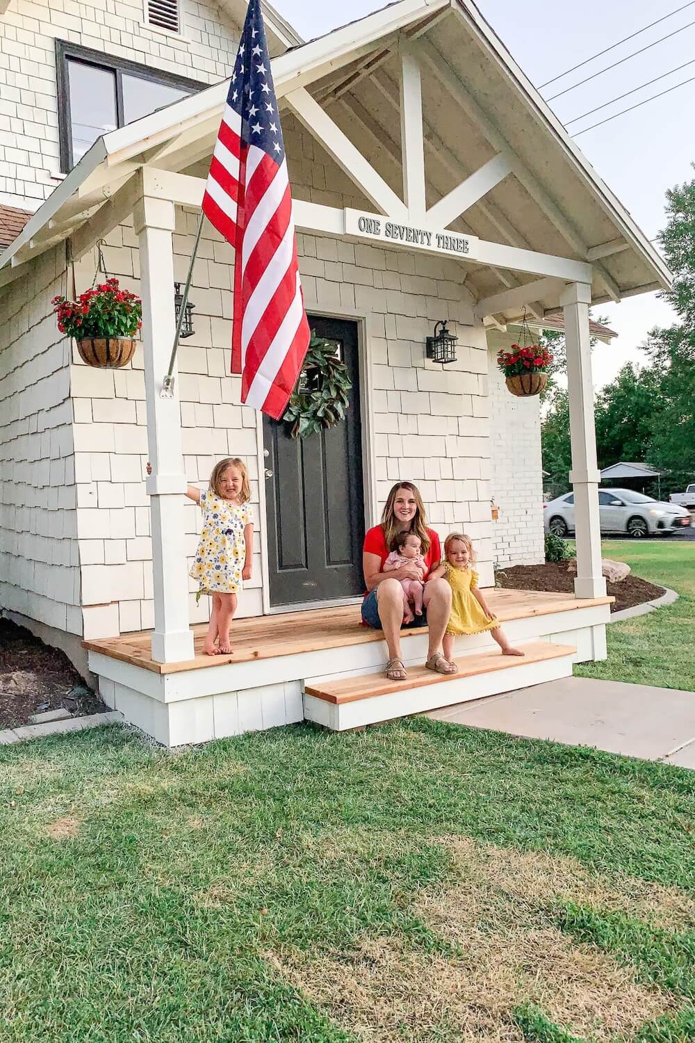 Woman and three young kids sitting on the steps of a finished DIY front porch with an American flag and magnolia wreath.
