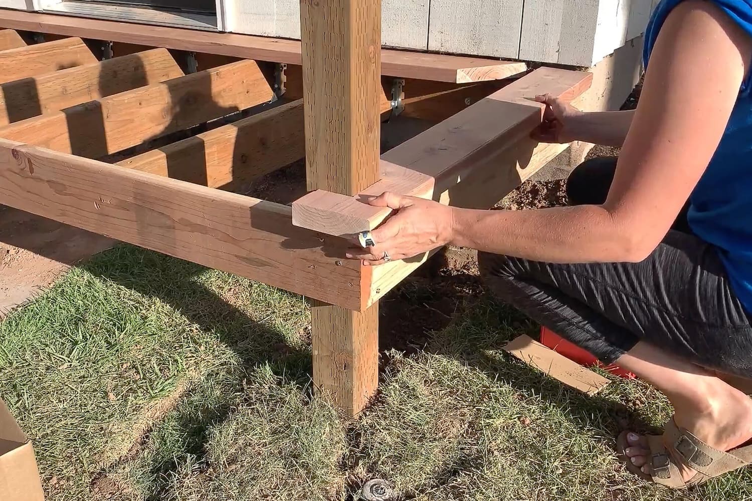 Woman fitting a notched redwood trim board snugly around a porch support post.