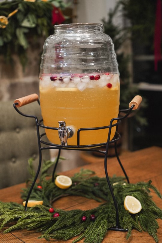 A large glass drink dispenser filled with copycat homemade Martinelli's, garnished with ice, lemon slices, and cranberries, sitting on a festive holiday table surrounded by greenery and lemons.