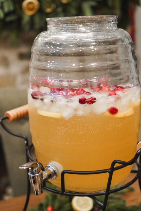 Close-up of a glass drink dispenser filled with copycat homemade Martinelli's, topped with ice, cranberries, and lemon slices, ready to be served for a holiday gathering.