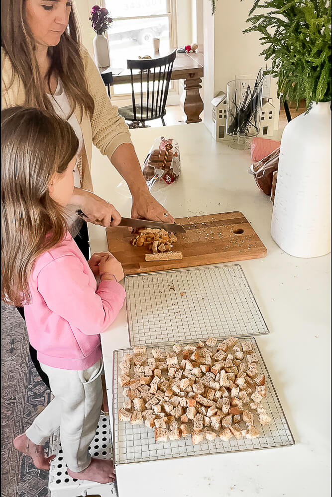 A mother and daughter chopping bread on a wooden cutting board, preparing bread cubes for homemade Thanksgiving stuffing.