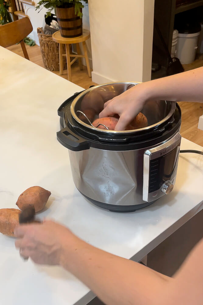 A person placing whole sweet potatoes into an Instant Pot for steaming, with the Instant Pot sitting on a kitchen countertop.