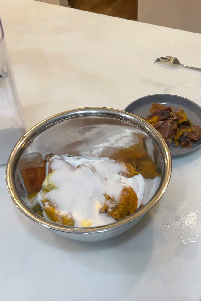 A metal mixing bowl containing cooked sweet potatoes, sugar, and butter, with sweet potato skins placed on a nearby plate.