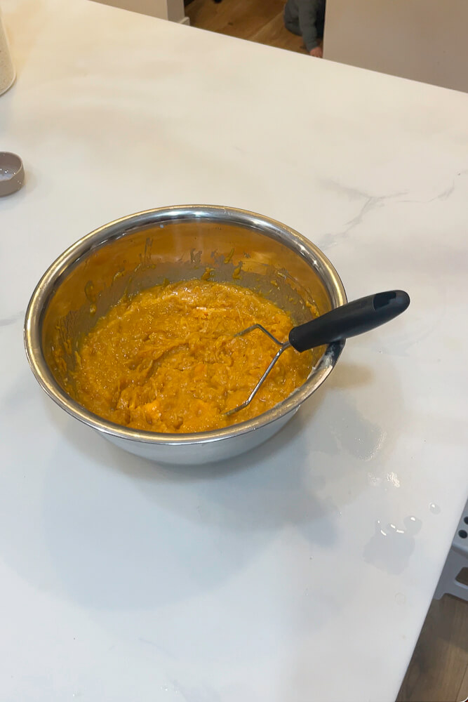 A metal mixing bowl filled with mashed sweet potatoes, ready to be spread into a casserole dish, with a potato masher resting in the bowl.