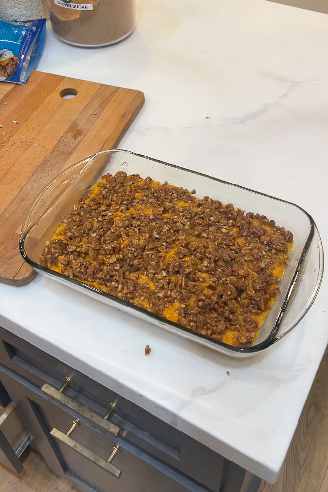 An unbaked sweet potato casserole topped with pecan crumble in a glass baking dish, placed on a kitchen countertop next to baking ingredients like brown sugar and shredded coconut.