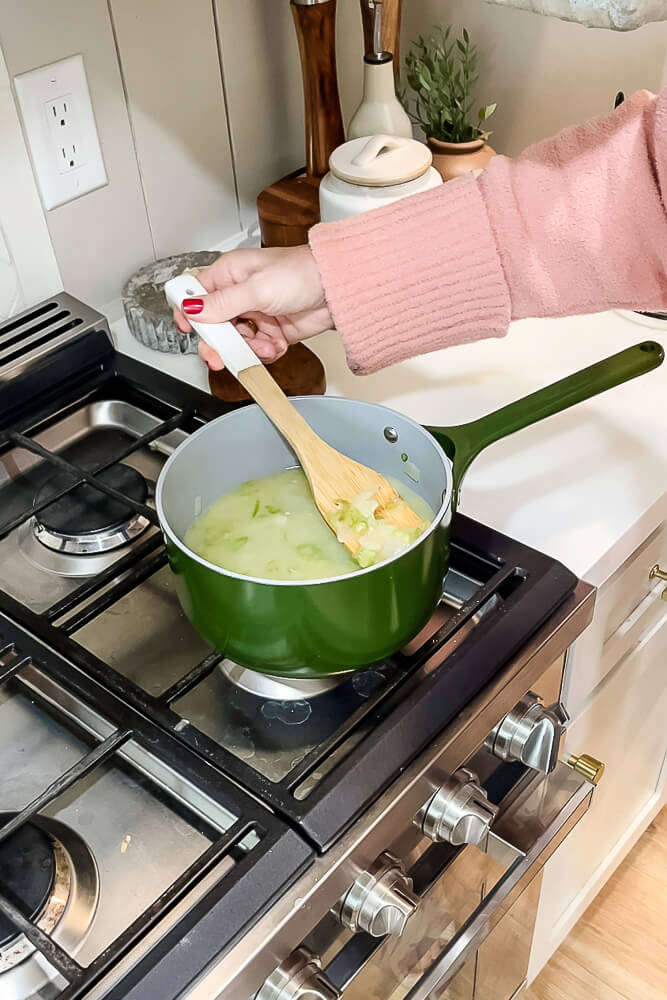 A wooden spoon stirring sautéed onions and celery in a green saucepan, preparing ingredients for Thanksgiving stuffing.