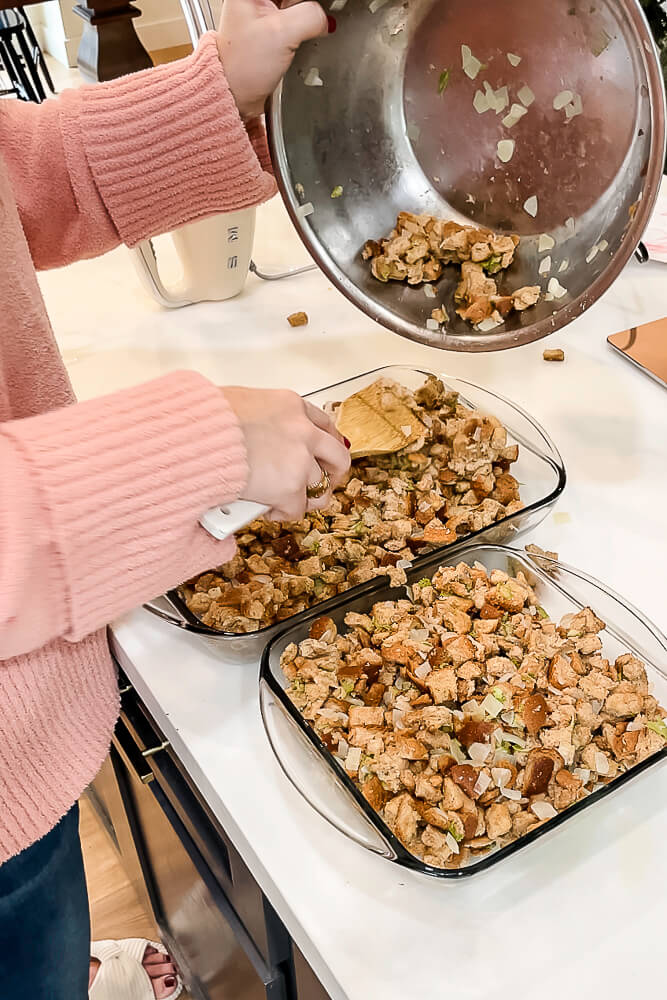 A person transferring homemade Thanksgiving stuffing from a large mixing bowl into two glass baking dishes, ready to be baked.