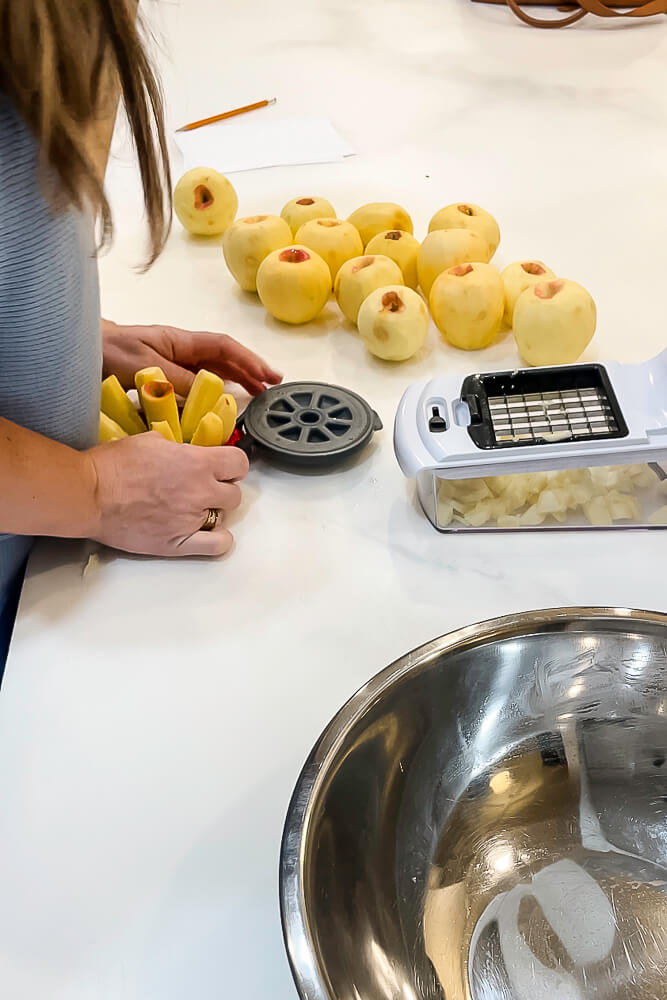 A person using a handheld apple slicer to slice peeled apples with another person assisting, and a bowl of chopped apples in the background.