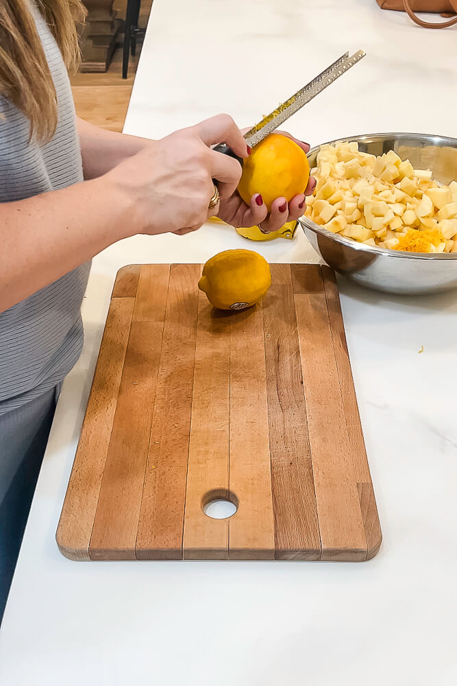 A close-up of hands zesting an orange over a wooden cutting board with a bowl of chopped apples in the background.