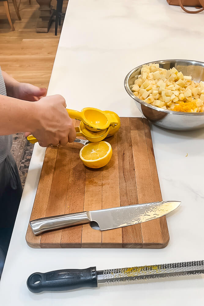 A person squeezing orange juice using a yellow citrus juicer over a wooden cutting board, with lemon halves and a large knife next to the cutting board.