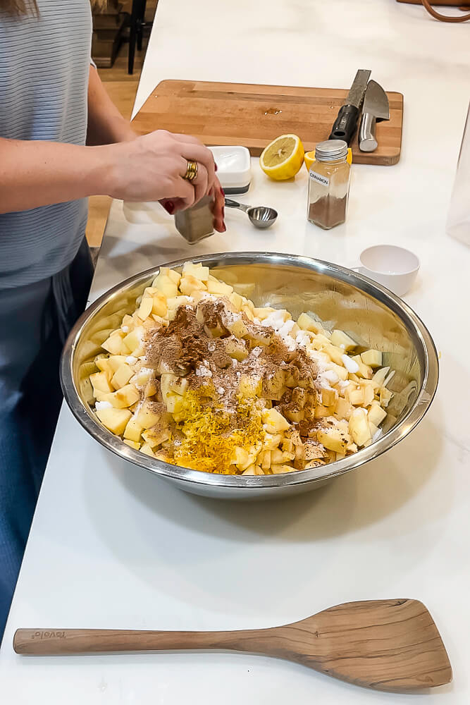 A large metal mixing bowl filled with chopped apples being seasoned with cinnamon, sugar, and orange zest, as someone sprinkles spices.