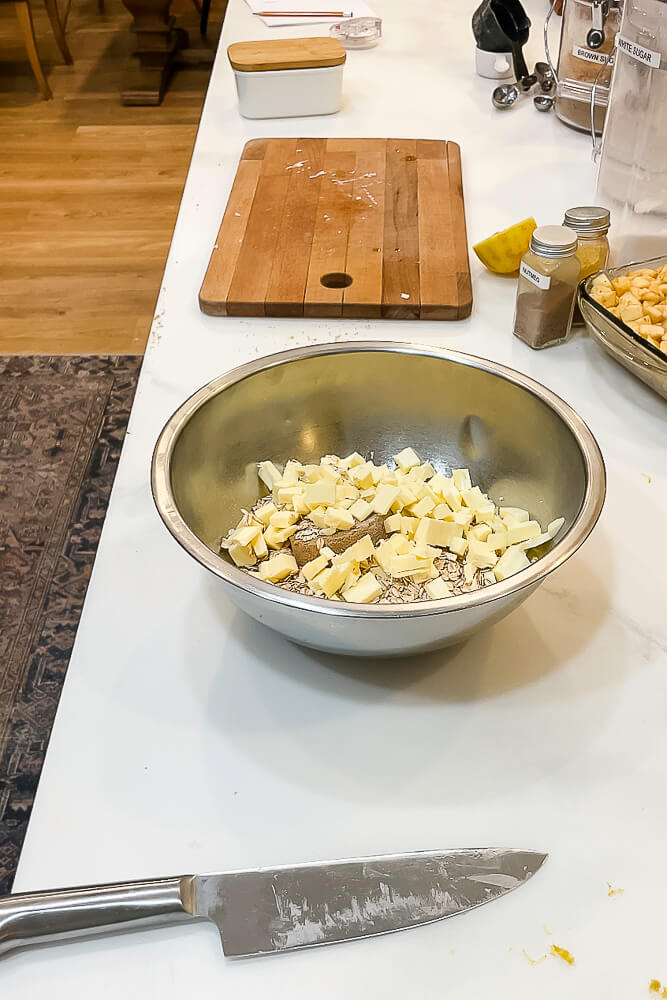 A mixing bowl filled with chopped butter and oats, ready to be combined into the crumble topping, with ingredients and a cutting board in the background.