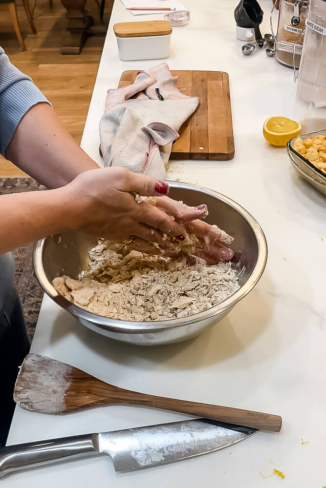 A person using their hands to mix together the ingredients for the crumble topping in a metal bowl, with a wooden spatula resting nearby.