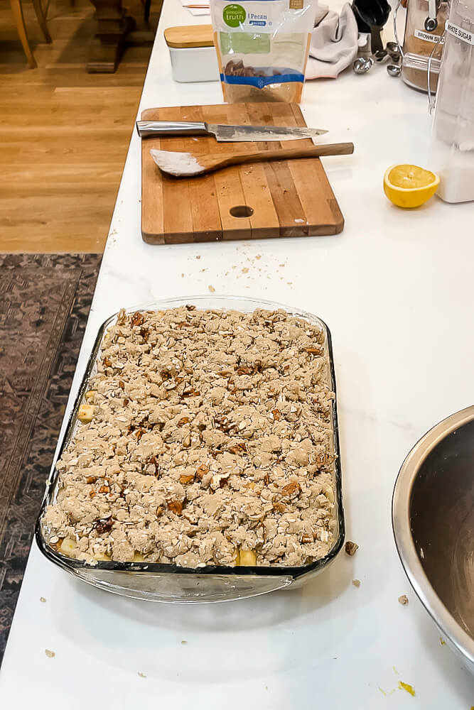 An assembled apple crisp topped with oat and pecan crumble, ready to be baked in a glass dish, sitting on a kitchen counter with a lemon half and utensils nearby.