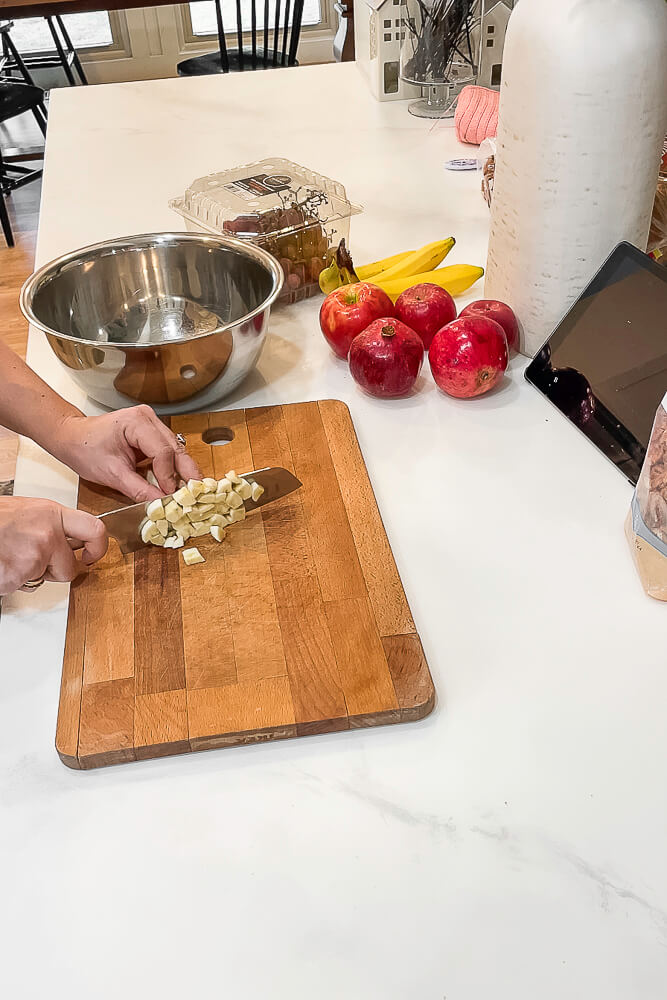 A hand is chopping small cubes of apples on a wooden cutting board with a stainless steel mixing bowl in the background.