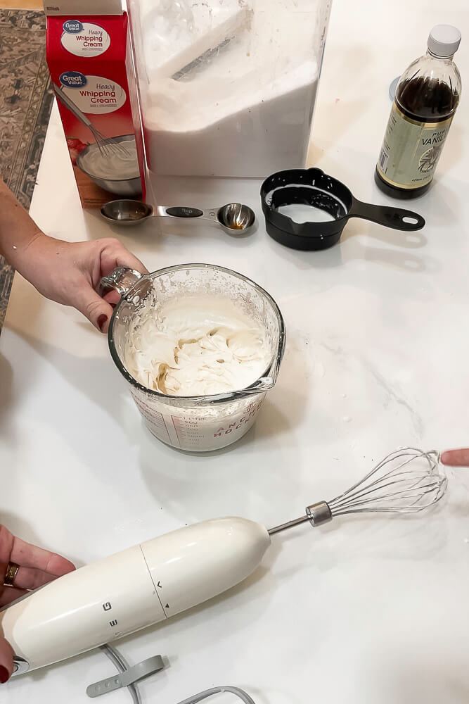 A close-up of an electric hand whisk blending cream in a glass measuring cup, with vanilla extract and other ingredients on the counter.