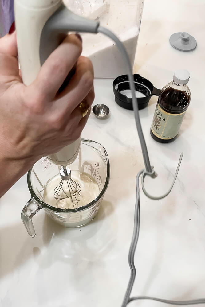 A hand-held mixer is used to whip heavy cream in a measuring cup on a white countertop, with ingredients like vanilla and sugar nearby.