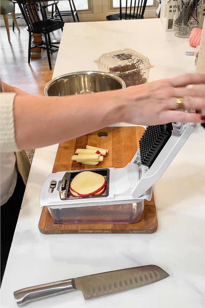 A hand is placing a red apple into a fruit dicer, with diced apple slices being collected into a clear container below.