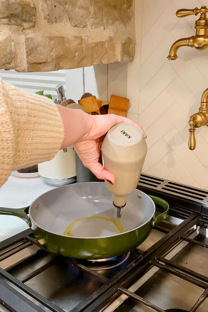 A hand drizzling oil into a green pan on the stove, preparing to sauté fresh green beans.