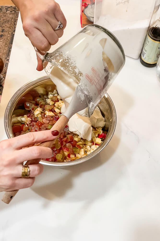 A spatula is used to scoop whipped cream into a mixing bowl filled with chopped apples and grapes, blending the salad ingredients.