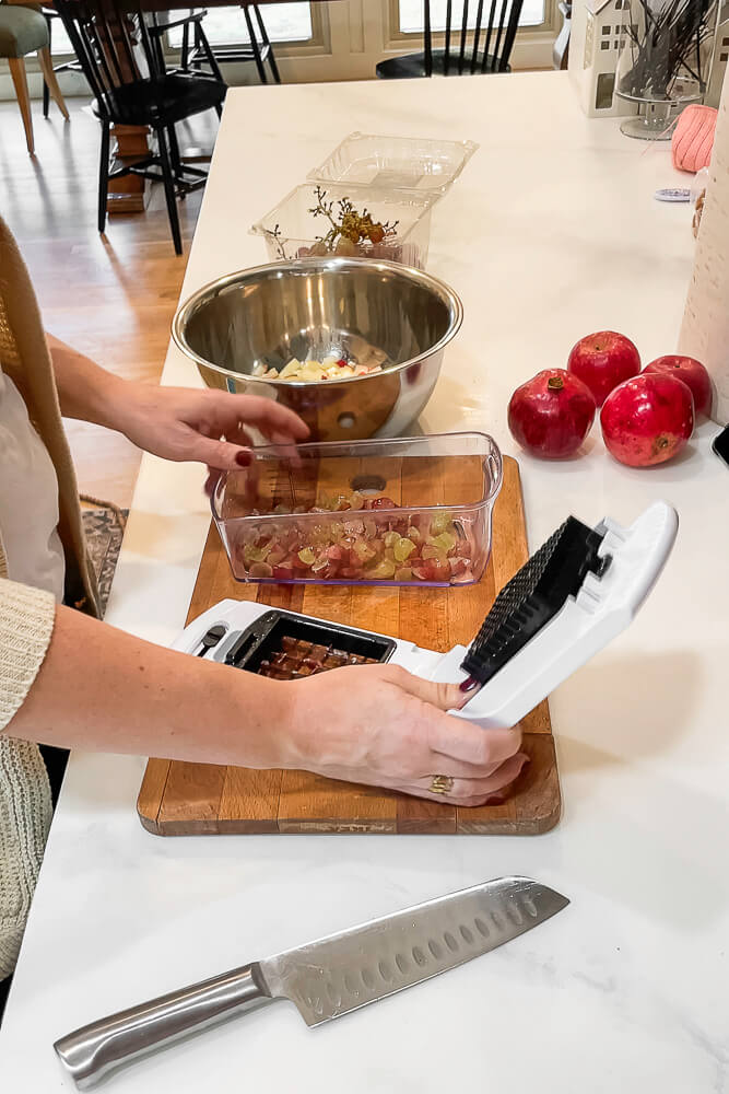 A hand operates a fruit dicer, cutting grapes into small pieces for the salad, with other ingredients on the countertop.