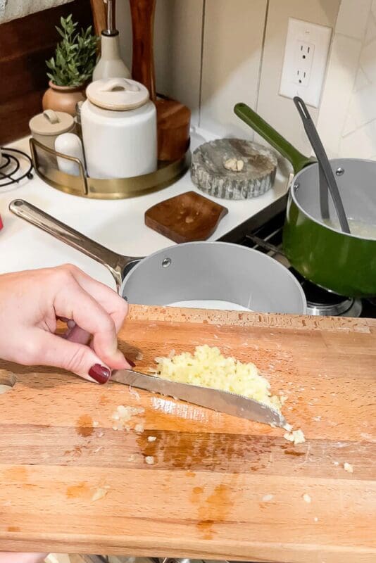 Minced garlic being scraped from a cutting board into a pot, adding flavor to garlic mashed potatoes