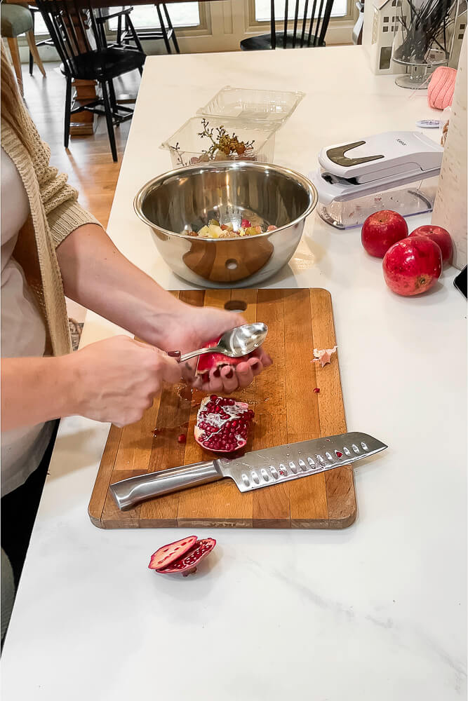 A hand uses a spoon to scoop out pomegranate seeds from a halved pomegranate, preparing ingredients for the salad.