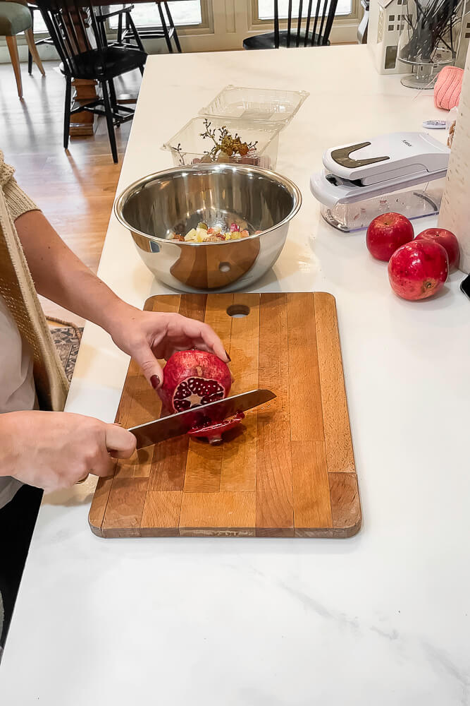 A hand holds a pomegranate as it is being sliced on a wooden cutting board, with a mixing bowl in the background.