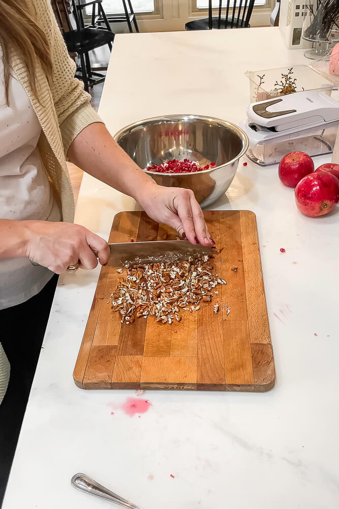 A person chopping pecans on a wooden cutting board, with a bowl of pomegranate seeds and whole pomegranates on a kitchen counter in the background.